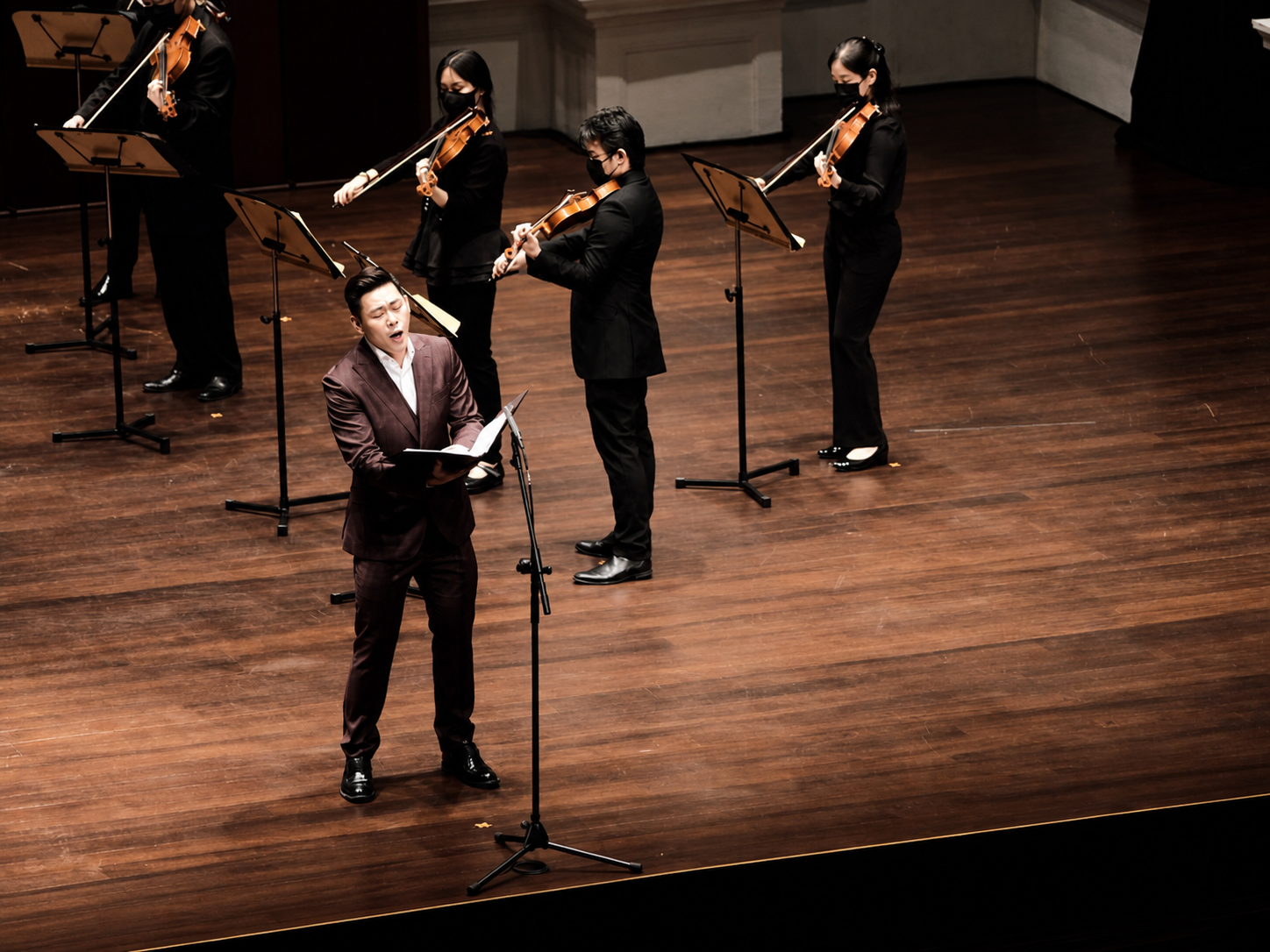 Alvin singing in concert with masked string ensemble in a wooden-floored hall