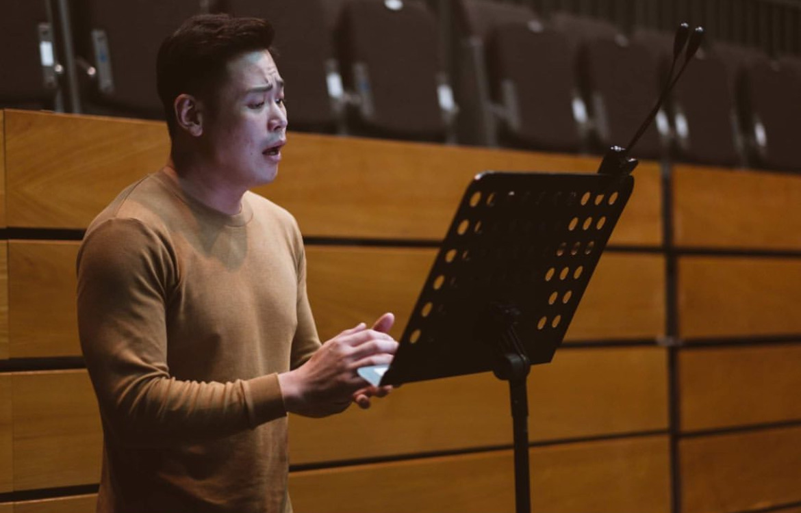 Alvin in a tan sweater rehearsing with a music stand in a wooden-panelled hall