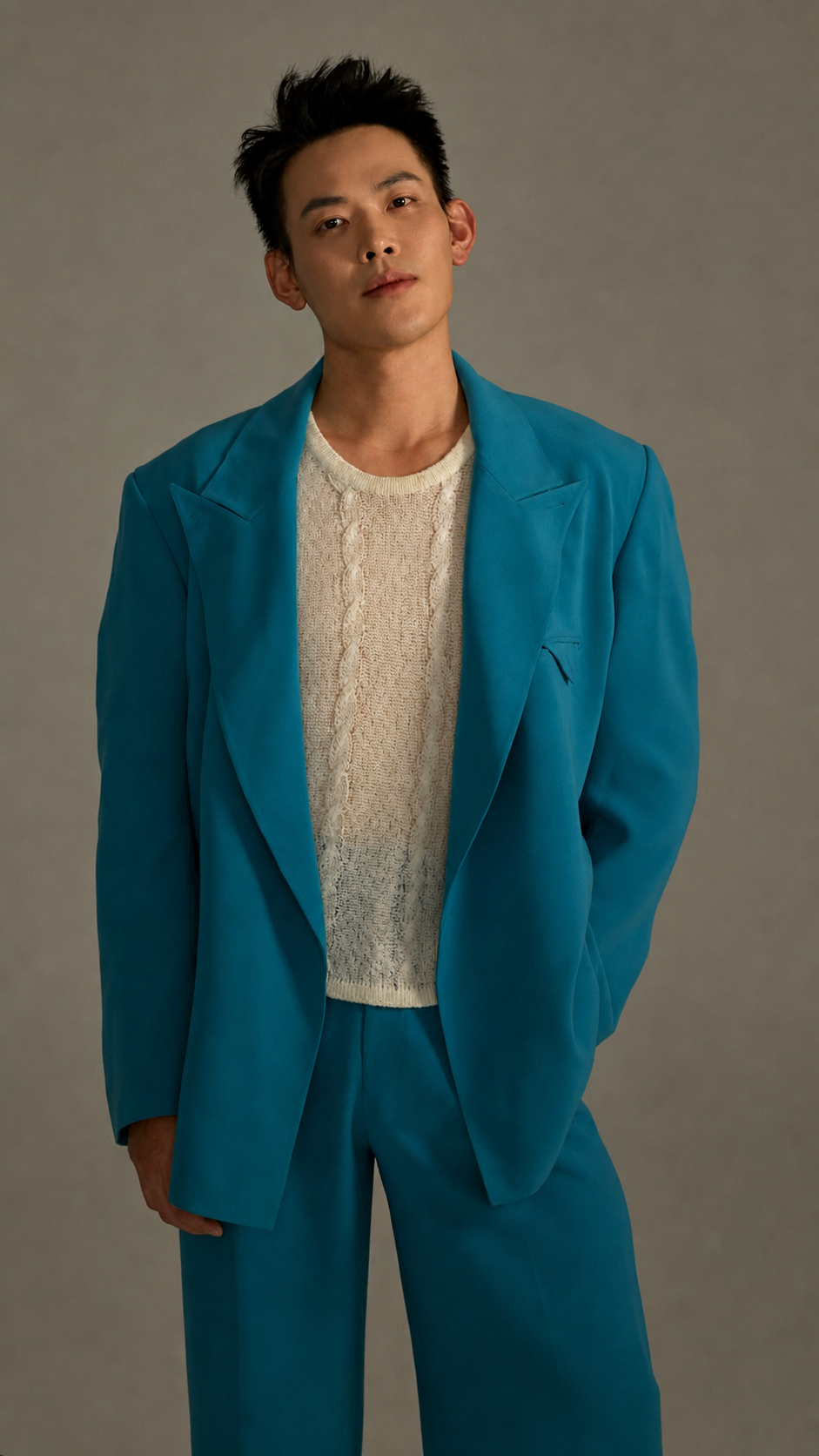 Editorial portrait. Alvin in oversized teal blazer over cream cable-knit sweater, on a neutral backdrop