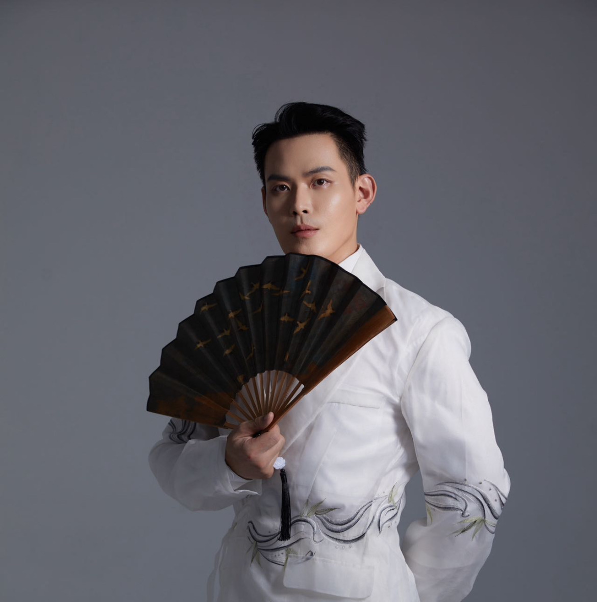 Alvin in a white embroidered jacket holding a black Chinese fan painted with golden birds in flight